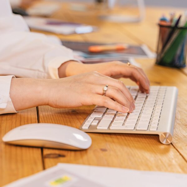 Slika prikazuje sto svetlo braon boje na kome je tastatura bele boje. Na slici su vidljive i zenske ruke kako kucaju na tastaturi,na stolu je i mis bele boje i kutija sa olovkama i casa sa vodom.